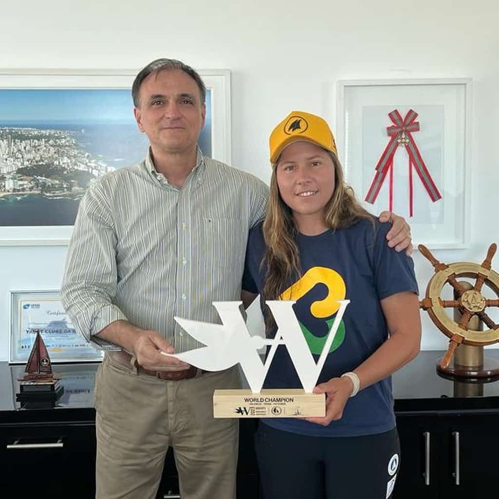 Na imagem, capturada da esquerda para a direita, temos o Comodoro do Yacht Clube da Bahia, Ricardo Dantas, acompanhado da atleta Juliana Duque. Ambos posam para a foto segurando um troféu com orgulho. Juliana Duque, usando um boné, veste uma camisa azul petróleo da Petrobahia, decorada com um coração no centro. A expressão de satisfação e conquista em seus rostos reflete a importância desse momento especial. A composição da imagem transmite a parceria entre o clube e a atleta, celebrando seu desempenho excepcional e a dedicação ao esporte.