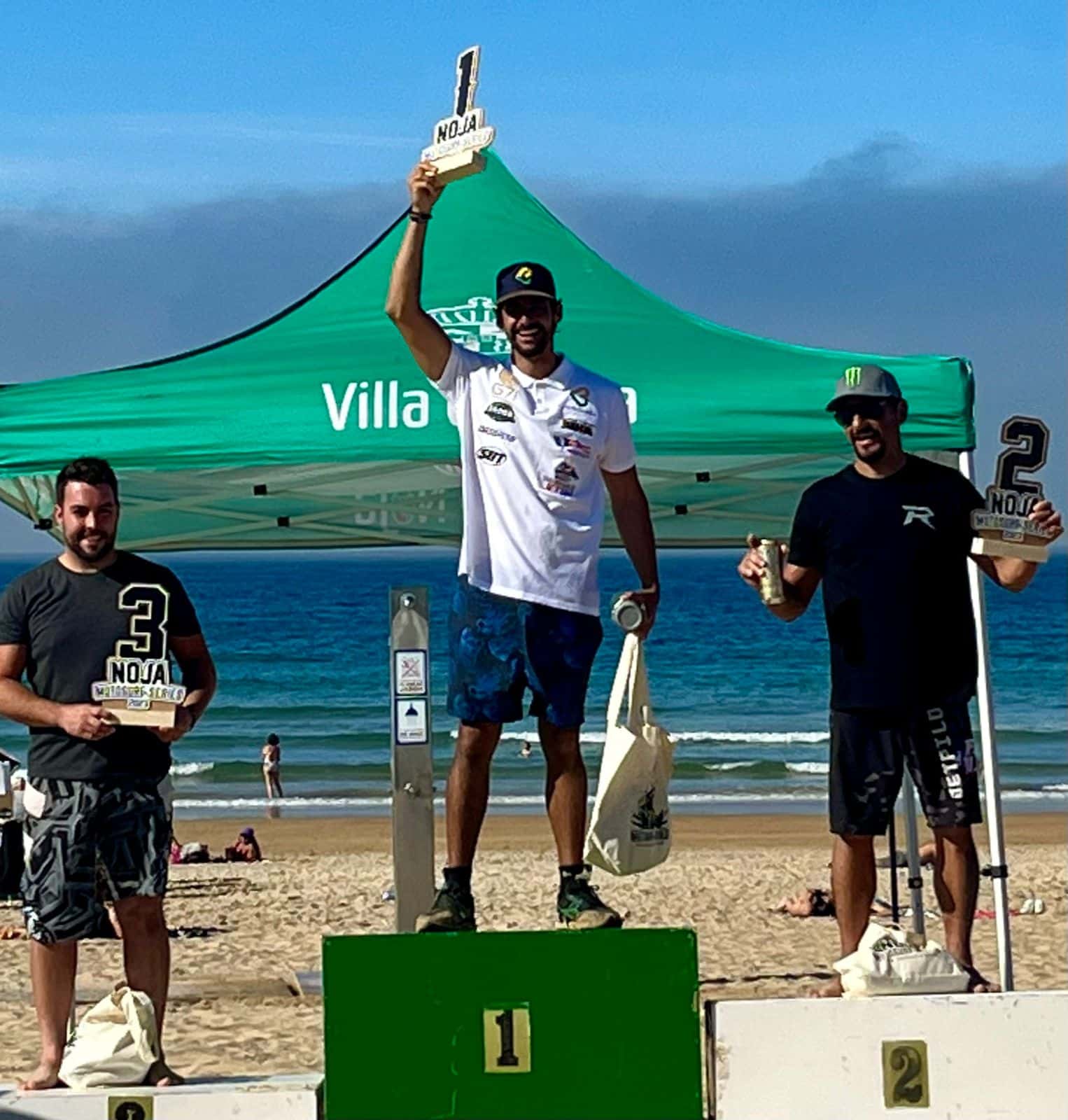 Na imagem vibrante capturada na praia, vemos o momento de glória do atleta Bruno Jacob, que ocupa o 1° lugar do pódio. Com um sorriso radiante, Bruno ergue com orgulho o troféu acima da cabeça, celebrando sua conquista. Sua aparência é evidente, vestindo uma camisa branca que realça sua determinação e uma bermuda azul que reflete sua confiança.