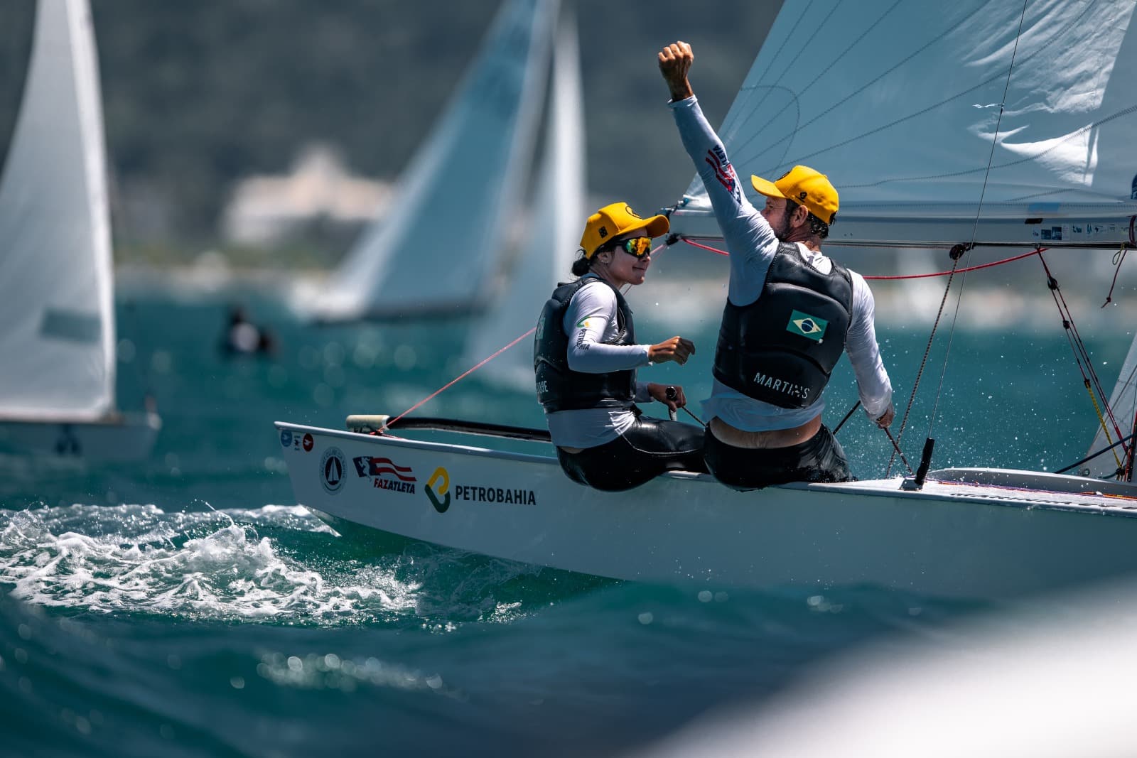 Na imagem , somos apresentados à atleta Juliana Duque e seu parceiro, ambos exibindo uma incrível habilidade de velejar em alto mar. Eles estão adequadamente equipados com coletes e bonés, demonstrando seu compromisso com a segurança e o conforto durante a prova. 